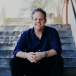 Image description: author Laurie Steed sits on steps. He is smiling at the camera and his arms rests on his knees.