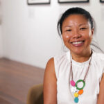 Image description: Artist Jacky Cheng is sitting in an art gallery. She is sitting to the right of the centre of the photo and is smiling into the camera. Photo credit: Amelia Blanco