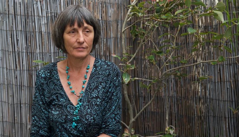 ID: Author Brigid Lowry sits in a garden with a bamboo fence behind her. She is looking off-camera in a contemplative mood.