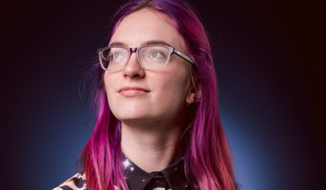 ID: A young woman with long pink hair and glasses glances upwards slightly in a contemplative mood.