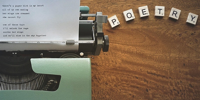 ID: on a wooden desk is an old-fashioned typewriter, with a poem typed up on paper. To the right of this are five scrabble pieces that spell out the word Poetry.