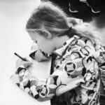 ID: black-wand-white photo of a young girl with long hair leaning over a desk with pencil in hand and writing on a p iece of paper.