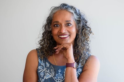 ID: a mid-shot of writer Asha Rajan, who is wearing a blue dress with a while floral pattern. Her chin rests on her hand and she is smiling directly into the camera.